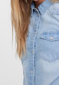 Camicia di denim azzurro chiaro con maniche lunghe, colletto a punta, due tasche frontali con patta e chiusura a bottoni metallici. Texture morbida e design casual.