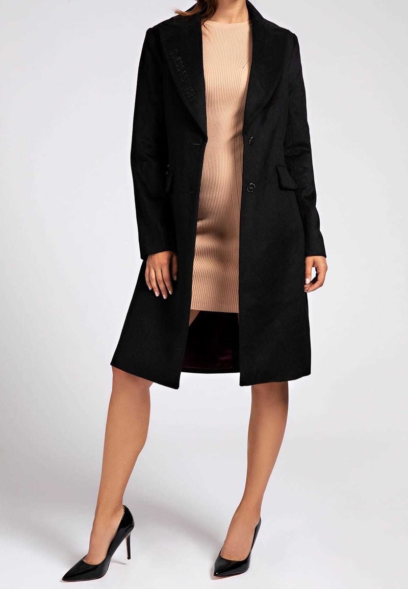 Zwarte wollen jas met een op maat gemaakt ontwerp, met notch-lapels, twee knopen en zijzakken, gecombineerd met een geribbelde beige jurk en zwarte hakken.