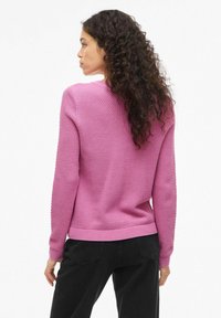 Różowy sweter dzianinowy o fakturze, z okrągłym dekoltem i ściągaczami na końcach rękawów. Noszony z czarnymi spodniami, prezentujący luźny krój i casualowy styl.