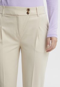Pantaloni beige su misura con pieghe, dotati di due bottoni marroni in vita e una superficie liscia. Tasche visibili sul lato.