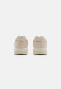 Paire de baskets beige New Balance vue de dos, mettant en avant le matériau en daim et le logo blanc brodé sur les languettes du talon.