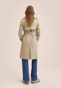 Béžový trenčkot s páskem v pase, bočními popruhy a zadním větráním. Volné modré džíny a šedé tenisky doplňují outfit.
