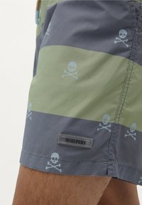Shorts de bain avec des rayures horizontales grises et vertes alternées, décorés de crânes et os croisés bleu clair, avec une étiquette "SCALPERS".