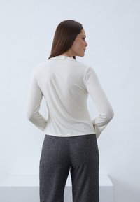 Langärmliges Creme-Oberteil mit gerippter Textur und Stehkragen, kombiniert mit grauen, strukturierten, weiten Hosen. Rückansicht dargestellt.