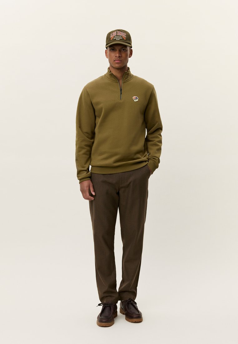 Sweat-shirt en polaire vert olive avec col zippé, patch logo. Pantalon à carreaux marron. Chaussures foncées. Le mannequin se tient devant un fond neutre.