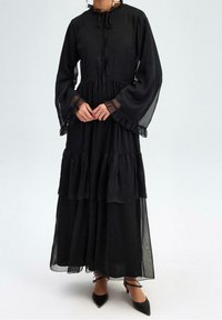Robe noire à manches longues à volants avec un col haut, des manches transparentes et des poignets à volants. Comprend un cordon à la nuque pour un réglage.
