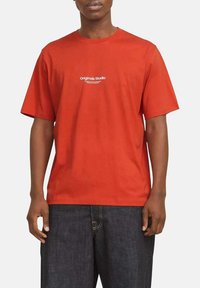 Rød bomulds T-shirt med rund halsudskæring, korte ærmer, med et hvidt logo trykt på brystet. Båret med mørkeblå denim bukser.