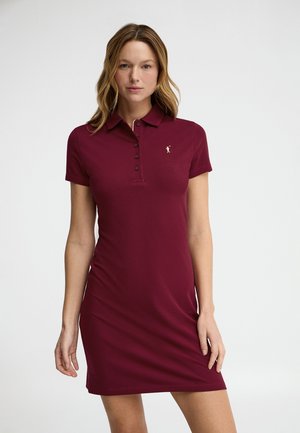 Frau trägt ein kurzärmliges, geknöpftes, burgunderrotes Polokleid mit kleinem Logo auf der Brust, steht vor einem einfarbigen Hintergrund.