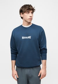 Νεαρός άνδρας που φοράει μπλε μπλουζάκι New Balance με μακριά μανίκια, κοιτάζοντας πλάγια σε ένα απλό ανοιχτόχρωμο φόντο.