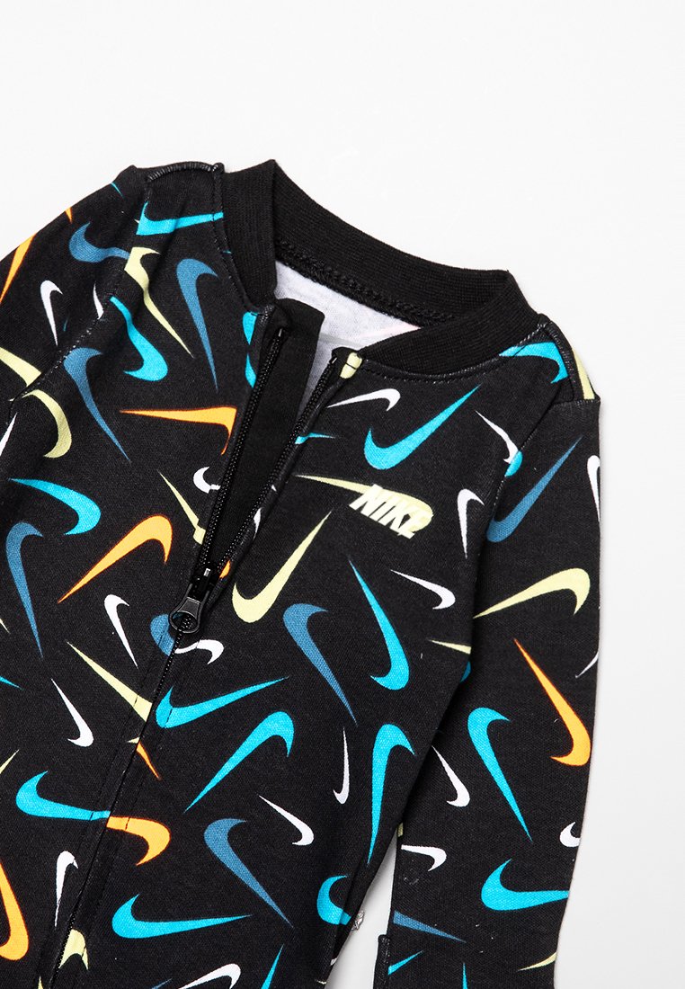 Combinaison noire zippée avec un motif de swoosh Nike multicolore en turquoise, orange, jaune et blanc. Tissu doux avec une coupe ajustée.