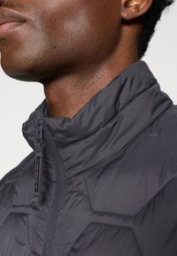 Close-up of a person wearing a dark quilted jacket with a high collar and visible zipper pull tab on the front.