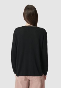 Vrouw met lang bruin haar, gekleed in een zwarte lange mouwtop en lichtroze broek, gezien van achteren tegen een effen grijze achtergrond.