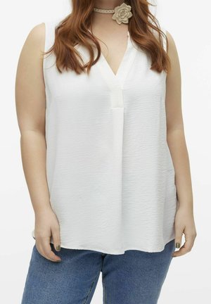 Femme portant un chemisier blanc texturé sans manches avec col en V et un collier ras du cou floral, assorti à un jean bleu, sur un fond uni.