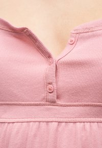 Κοντινή λήψη ενός ροζ ριμπ πλεκτού τοπ με τρία μικρά κουμπιά, το ένα μερικώς ξεκουμπωμένο, που αποκαλύπτει μια σεμνή γραμμή λαιμού.