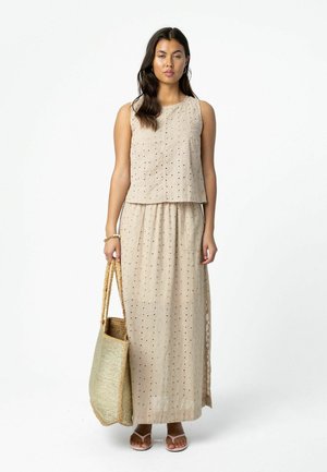 Naine, kes kannab varrukateta beeži silmkoe topi ja sobivat pikkade seelikuga, hoides suurt punutud käekotti, seisab valge tausta ees.