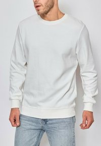 Sweatshirt en coton blanc avec une texture côtelée, encolure ras du cou et manches longues. Ourlet et poignets ajustés dans une couleur blanche assortie.