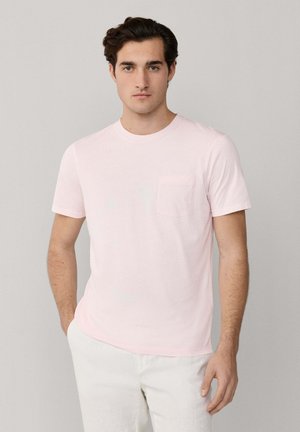 Young man wearing a light pink short-sleeve T-shirt with a chest pocket and white pants, standing against a plain gray background.