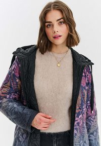 Mladá žena v květovaném prošívaném kabátu přes béžový pletený svetr, s krátkými hnědými vlasy a zlatým náhrdelníkem.