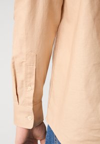Chemise beige à manches longues avec des poignets structurés, ornés de deux boutons et d'une texture lisse et propre. Hem et détails de coupe visibles.