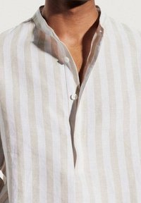 Camicia in lino leggero a righe orizzontali bianche e beige. Caratterizzata da un colletto alla mandarino e una pattina con bottoni frontale con un solo bottone.