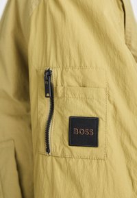 Žlutá lehká bunda s texturovaným povrchem, obsahující boční kapsu na zip a obdélníkovou černou nášivku s nápisem "BOSS" v hnědé barvě.