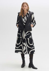 Schwarze Pufferweste mit Kapuze über einem schwarz-weiß gemusterten Kleid. Das Kleid hat einen fließenden Stoff und ein gestuften Design. Schwarze Stiefeletten werden getragen.