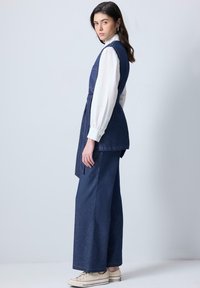 Une combinaison en denim avec des pantalons à jambes larges, une taille ceinturée et deux poches avant. Les longues manches blanches contrastent avec le tissu bleu foncé.