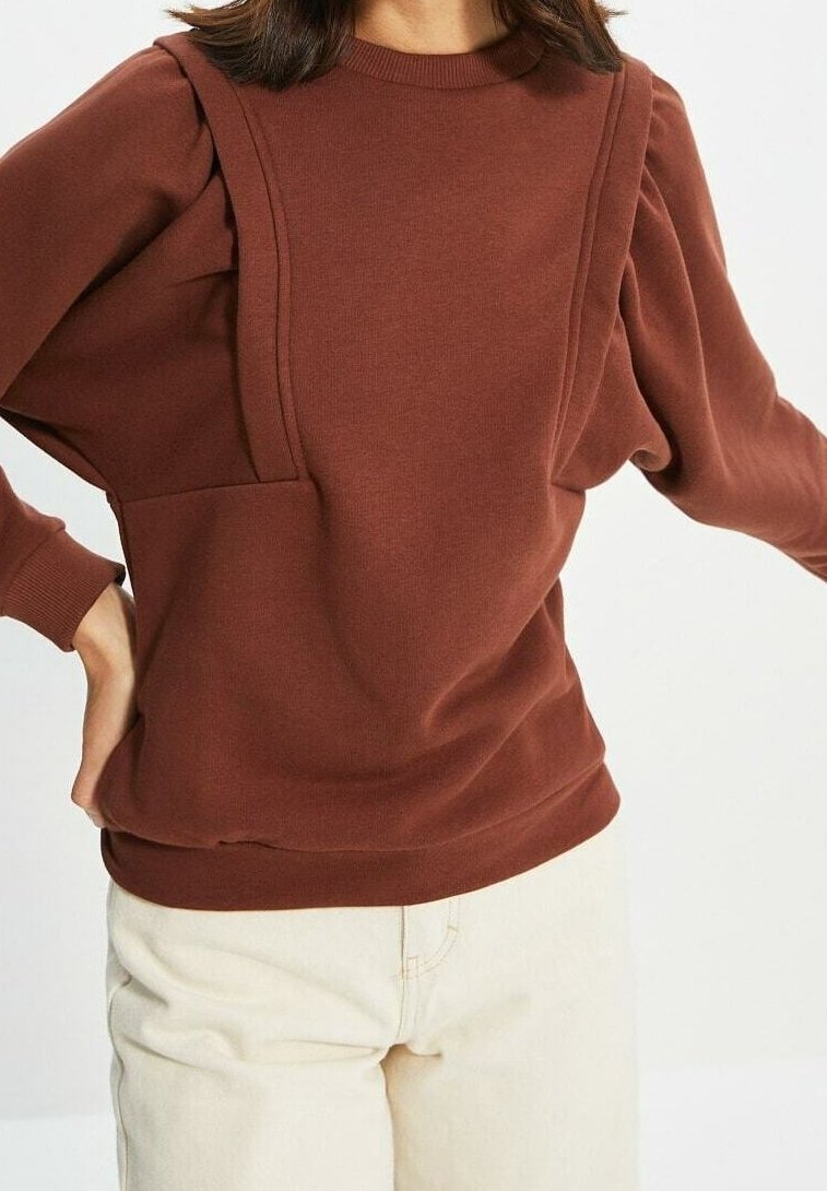 Sweatshirt marron avec manches bouffantes texturées, encolure côtelée et poche avant. Tissu doux et coupe décontractée ; associé à un pantalon de couleur claire.