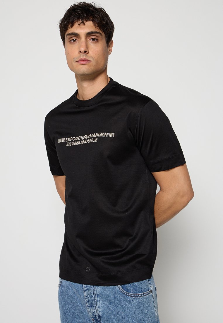 Hombre vestido con camiseta negra con el logo de Emporio Armani Milano y vaqueros azules, de pie con las manos detrás de la espalda contra un fondo liso.