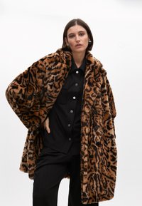 Kaput od umjetnog krzna s leopard uzorkom, širokim ovratnikom, slobodnim krojem i crnim gumbom, u kombinaciji s crnom košuljom na gumb i hlačama.