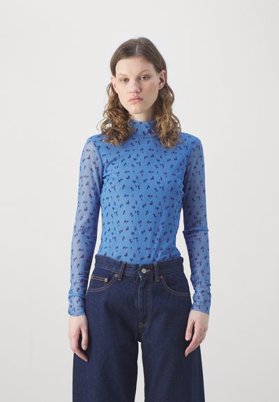 Top lungo a maniche trasparenti di colore azzurro chiaro con un motivo floreale in blu scuro e viola, abbinato a jeans in denim scuro a vita alta.
