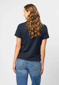 Kvinne med langt, bølgete brunt hår som har på seg en svart topp med korte ermer og blå jeans, vist bakfra mot en hvit bakgrunn.