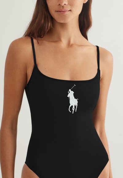 Μαύρο μαγιό με λεπτές τιράντες, με λευκό κέντημα λογότυπο αλόγου. Λείο υλικό, σχεδιασμός με ψηλή γραμμή που αναδεικνύει τη σιλουέτα.