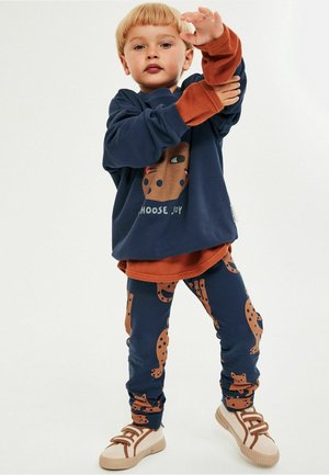 Marineblå sweatshirt med et brunt grafisk ansigtsdesign og tekst, kombineret med mønstrede marineblå leggings med brune katteillustrationer. Beige sneakers.