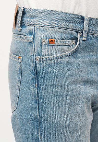 Približan posnetek svetlo modrih kavbojk z žepi spredaj in zadaj, oranžnimi oznakami ter vidnimi zankami za pas na osebi, ki nosi belo srajco.