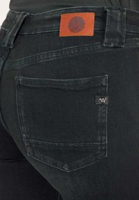 Jeans de mezclilla negra con una textura deslavada. Las características incluyen un bolsillo trasero rectangular, una pequeña etiqueta de marca y un parche de cuero en la cintura.