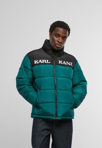 Kurtka pikowana w kolorze morskim i czarnym z wysokim kołnierzem, z wyraźnym białym napisem "KARL KANI" na piersi oraz zamkiem błyskawicznym.