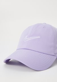 Lilla bomuldske med en buet skygge, med et hvidt broderet Nike-logo foran. Glat tekstur og struktureret design.