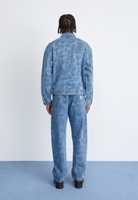 Blå denimjakke og bukser med et tonalt abstrakt mønster. Løs pasform med standardlommer og knapdetaljer. Sorte fodtøj.