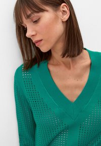 Groene gebreide trui met een V-halsontwerp. Heeft een opengeweven patroon langs de mouwen en het lichaam, met een gladde textuur en ribgebreide accenten.