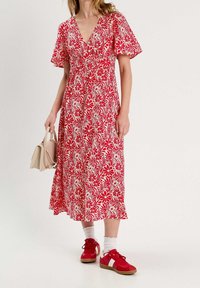 Robe midi à fleurs rouges avec des manches courtes, un décolleté en V et une ceinture à la taille. Assortie à des baskets rouges et un petit sac à main beige.