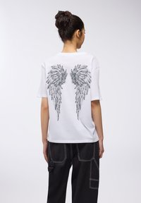 T-shirt bianco a maniche corte con un dettagliato design di ali nere stampato sul retro. Abbinato a pantaloni neri a gamba larga.