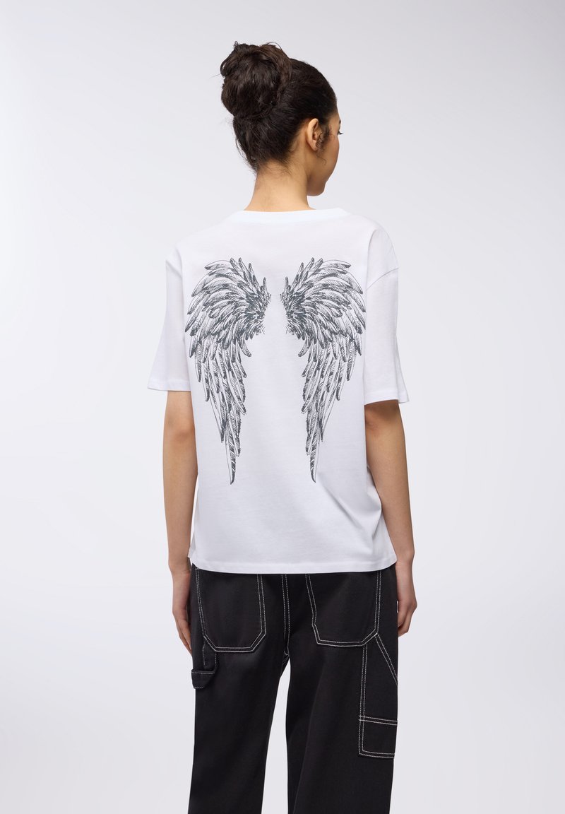 T-shirt bianco a maniche corte con un dettagliato design di ali nere stampato sul retro. Abbinato a pantaloni neri a gamba larga.