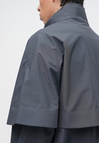 Tmavosivá bunda s vysokým golierom, štruktúrovanými ramenami a hladkou, matnou textúrou. Dizajn je moderný s čistou siluetou.