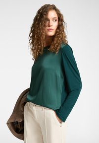 Vihreä pitkähihaine paita, jossa on sileä rakenne, pyöreä kaula-aukko ja sivutasku. Yhdistetty beigeihin housuihin ja ruskeaan takkiin.