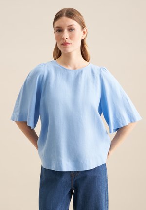 Frau mit hellbraunen Haaren, die eine lockere hellblaue Kurzarmbluse und dunkelblaue Jeans trägt, steht mit den Händen hinter dem Rücken.