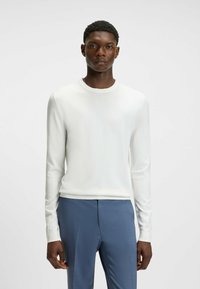 Camicia bianca a maniche lunghe con scollatura tonda, dalla texture morbida, abbinata a pantaloni blu su misura con un motivo discreto e pieghe frontali.