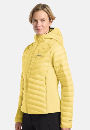 Eine Frau trägt eine gelbe gesteppte Jacke mit Kapuze und Reißverschluss, steht vor einem einfarbigen hellen Hintergrund und dreht sich leicht zur Seite.