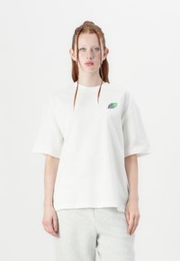 T-shirt surdimensionné blanc en coton, avec un petit logo vert et noir sur la poitrine gauche. Manches courtes et coupe décontractée.
