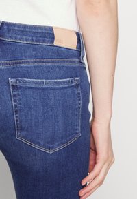 Tummansinisiä farkkuja, joissa on yksi takatasku ja beiget brändätyt nahkapatja vyötäröllä. Silkkinen rakenne, istuva muotoilu.